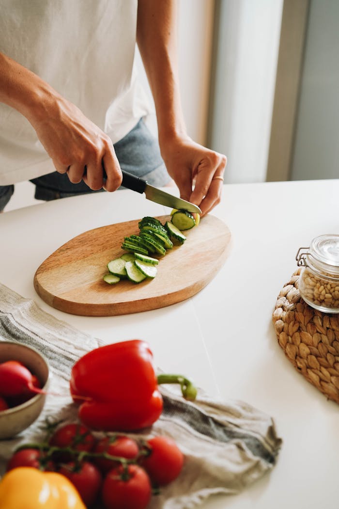 Close-up of hands slicing cucumber on a cutting board with additional fresh vegetables nearby.